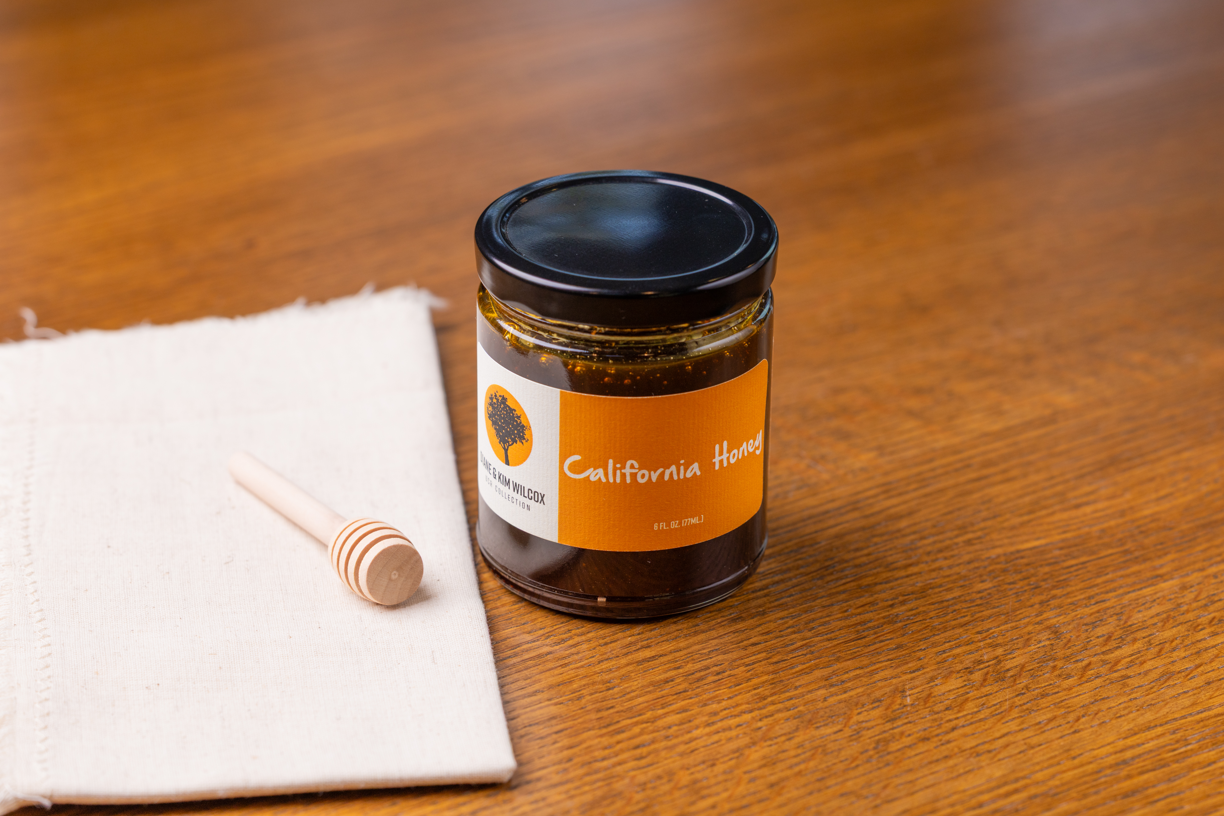 California Honey jar sitting on a wooden table with a cloth and honey stirrer.